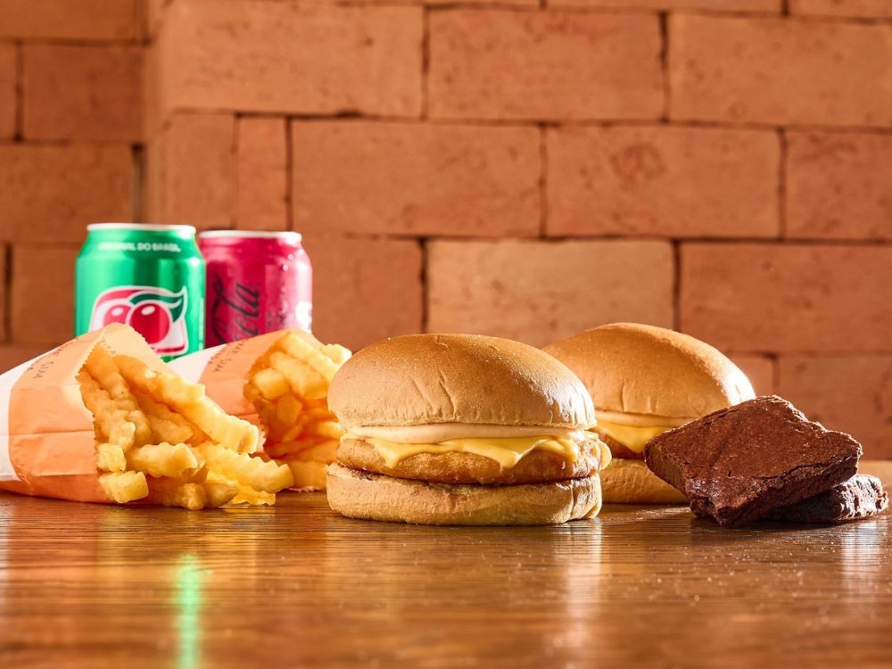 De que pais veio o hambúrguer? Leia e confira essa combinação de hambúrgueres, batata frita crocante, brownie e refrigerantes em mesa de madeira.