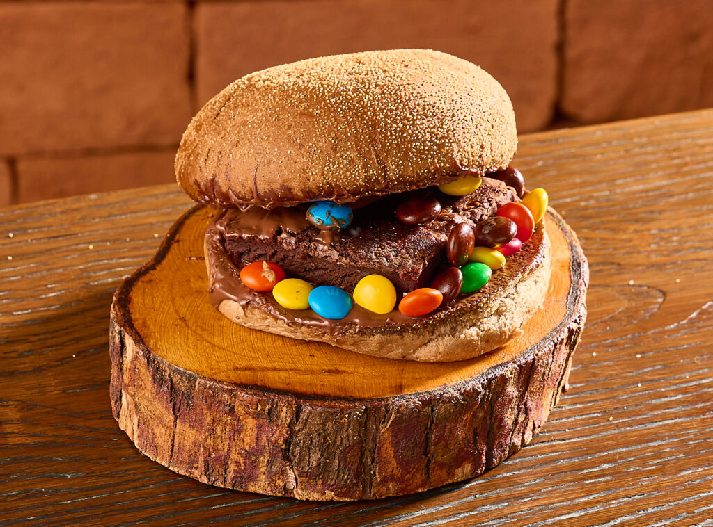Hambúrguer doce servido em pão com recheio de brownie, chocolate derretido e confeitos coloridos de chocolate, sobre uma base de madeira rústica.