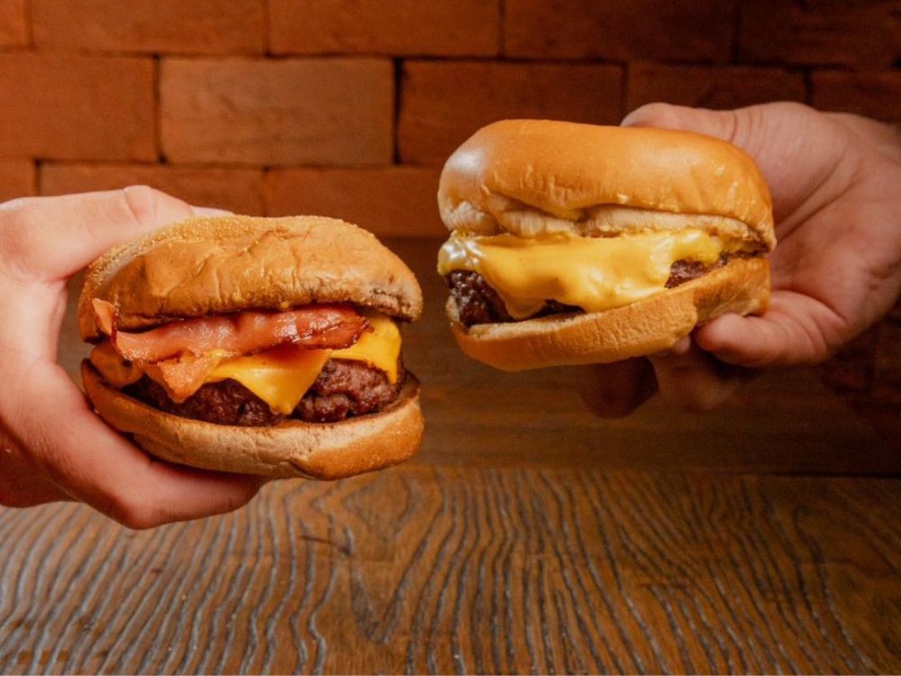 Duas mãos segurando hambúrgueres artesanais, um com bacon e queijo derretido e outro apenas com carne e queijo.