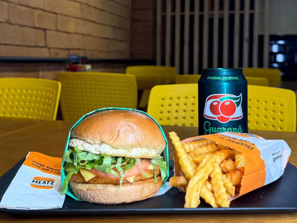 Combo com hambúrguer de frango empanado, alface, tomate e queijo, acompanhado de batata frita e lata de Guaraná, em mesa com cadeiras amarelas.