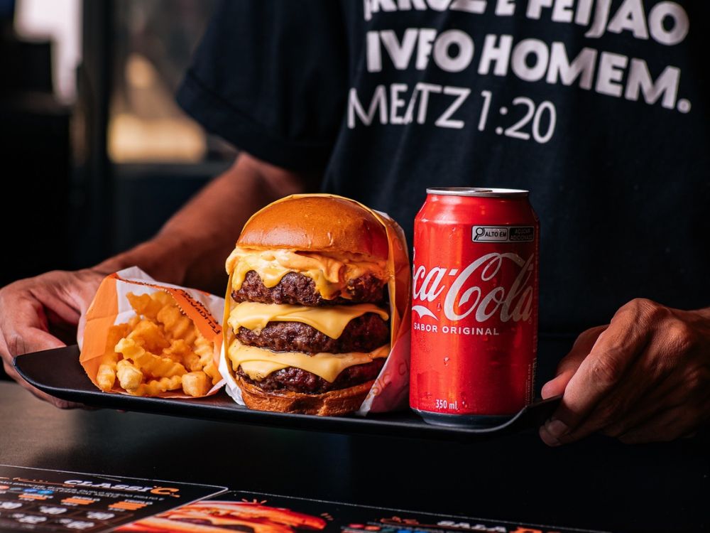 Bandeja com hambúrguer triplo de carne e queijo, batata frita crocante e lata de Coca-Cola sendo segurada por uma pessoa com camiseta preta.