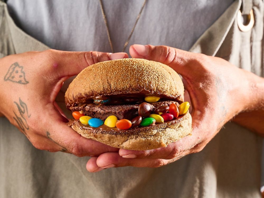 Pão artesanal recheado com chocolate, brownie e confeitos coloridos, mostrando a criatividade e diversidade na história do hambúrguer.