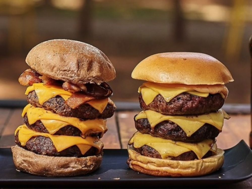 Dois hambúrgueres altos com camadas de carne e queijo, retratando a influência americana e a expansão mundial na história do hambúrguer.