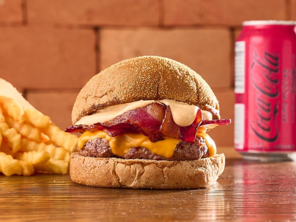 Hambúrguer artesanal com bacon e queijo ao lado de batatas fritas e refrigerante sobre a mesa.