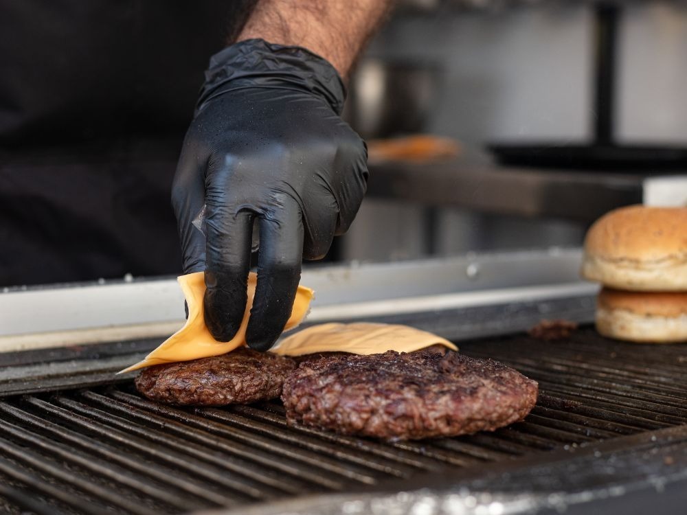 Mão com luva preta colocando fatia de queijo sobre hambúrgueres na chapa durante o preparo.
