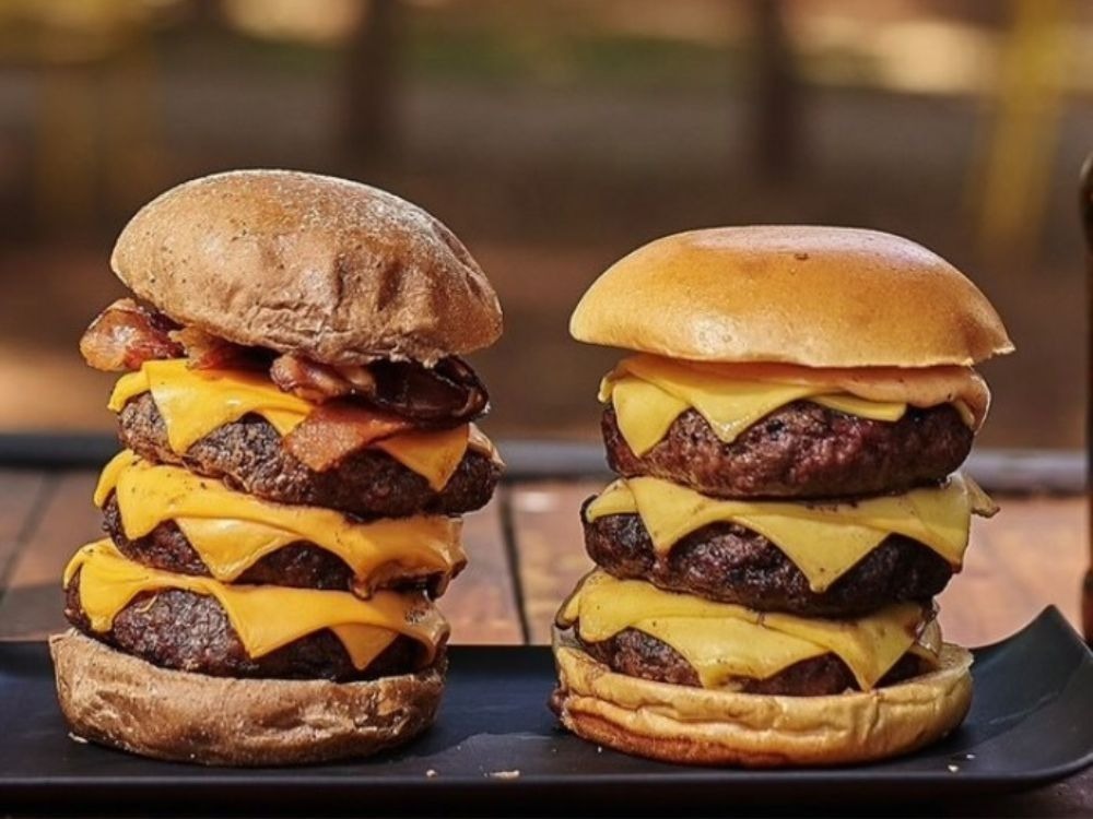 Dois hambúrgueres altos e suculentos com queijo e bacon, simbolizando o padrão de qualidade e sabor de uma franquia de hambúrguer artesanal.