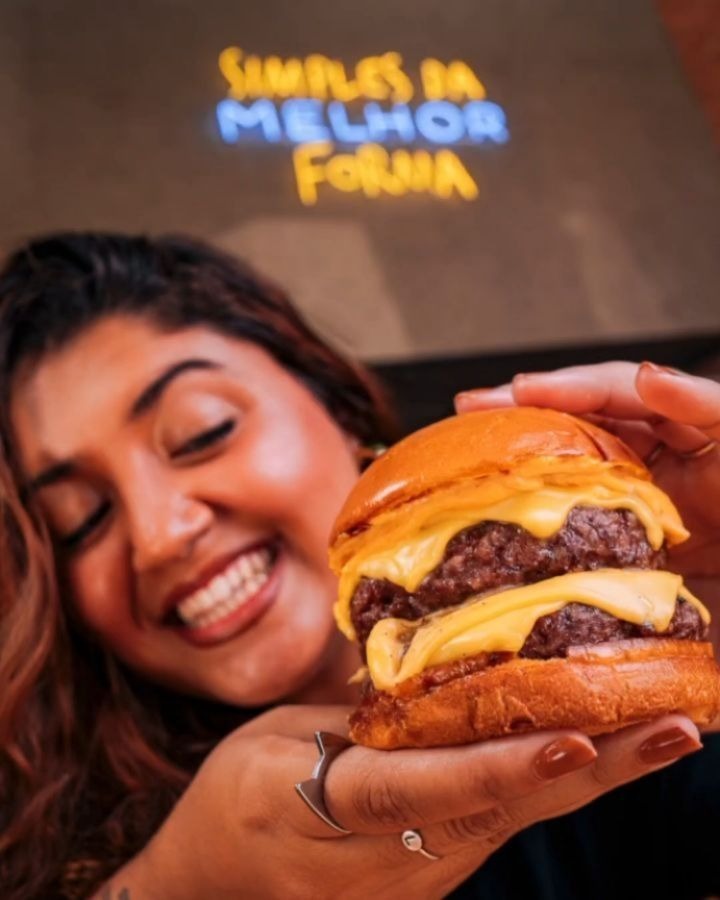 Cliente sorridente segurando hambúrguer artesanal duplo com queijo derretido, destacando a experiência saborosa em uma franquia de hambúrguer moderna.