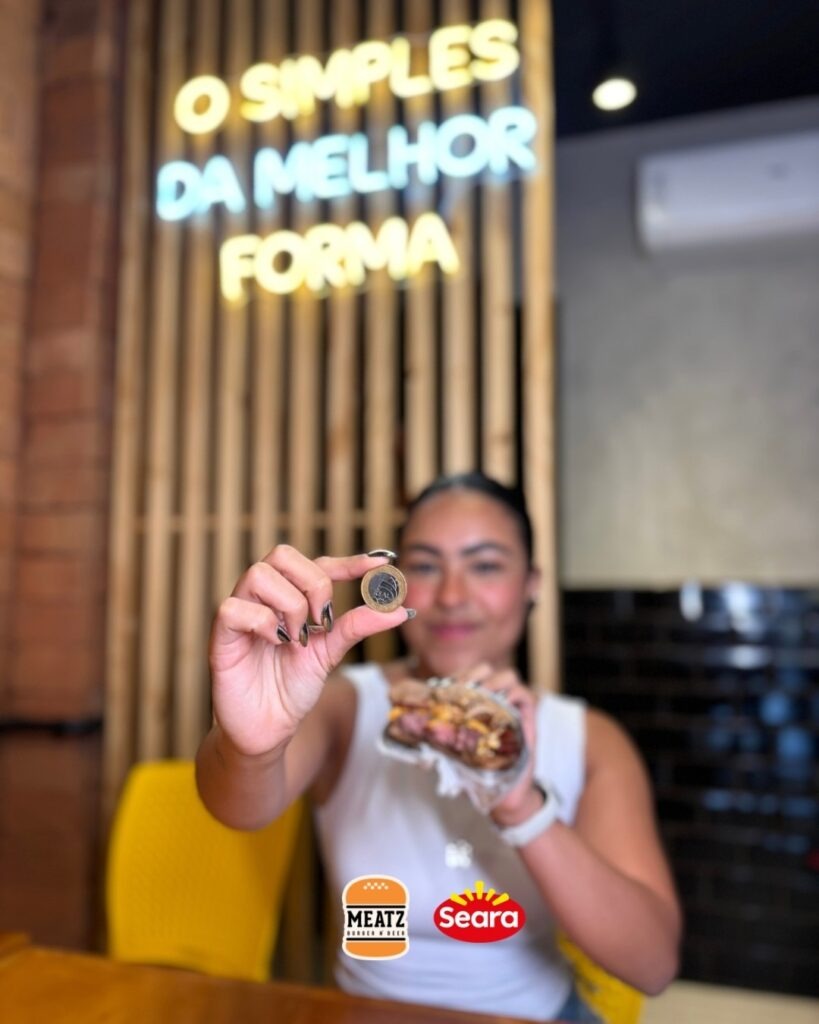 Pessoa segurando uma moeda enquanto mostra um hambúrguer em ambiente da Meatz Burger com letreiro iluminado ao fundo.