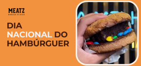 Hambúrguer doce com brownie e confeitos coloridos sendo segurado na mão ao lado do texto “Dia Nacional do Hambúrguer” da marca Meatz Burger n’ Beer em fundo laranja.